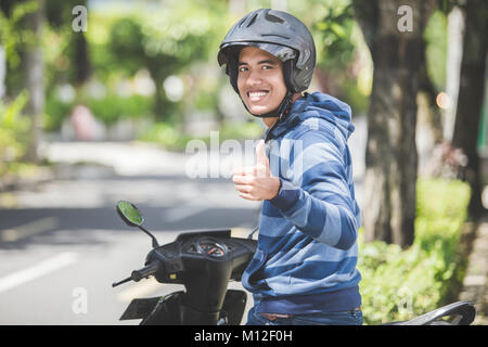 Ritratto di felice uomo asiatico a cavallo di moto in una strada di città e mostrando il pollice in alto Foto Stock