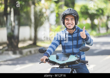 Ritratto di felice uomo asiatico a cavallo di moto in una strada di città e mostrando il pollice in alto Foto Stock