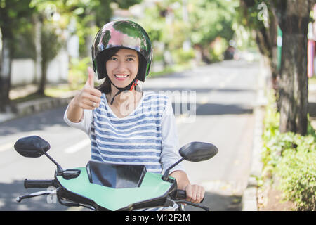 Ritratto di felice donna asiatica a cavallo di moto in una strada di città e mostrando il pollice in alto Foto Stock