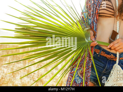 Bohemian vibe vacanza. Primo piano sul grande verde foglia tropicale in mano del bohemian chic di jeans corti e capo all'aperto in serata estiva Foto Stock