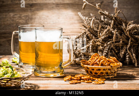 Due boccali di birra di schiuma stare in piedi sul tavolo di legno con salatini, frumento e luppolo Foto Stock