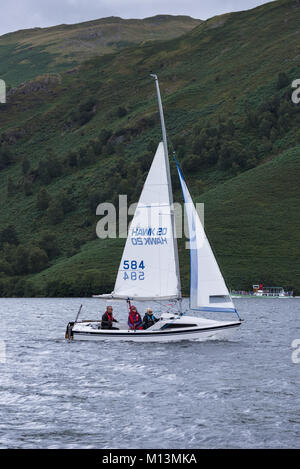 Sotto ripide colline, 3 persone sono in barca a vela nella piccola barca con 2 vele, sulle acque del lago, Ullswater vaporizzatore al di là - Lake District, Inghilterra, Regno Unito. Foto Stock