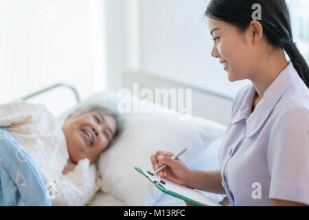 Infermiera asiatica a controllare la storia della malattia paziente anziano giacente in letto. Un infermiere si prende cura di una donna anziana. Foto Stock