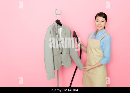 Professional giovani femmine servizio lavanderia Negozio dipendente utilizzando aspirapolvere pulizia attrezzo suit abbigliamento e in piedi di sfondo rosa guardando la telecamera sm Foto Stock