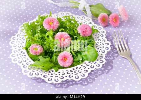 Piastra bianca con una insalata verde mista le foglie di lattuga e commestibili fiori a margherita, insieme con una forcella di vintage su un nostalgico sfondo lilla Foto Stock