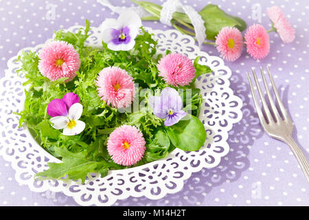 Piastra bianca con una insalata verde mista le foglie di lattuga e commestibile daisy e pansy fiori, insieme con una forcella di vintage su un nostalgico backgroun lilla Foto Stock