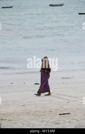 Donna musulmana passeggiate sulla spiaggia di Nungwi, Zanzibar, coperte dalla testa ai piedi e indossa una sciarpa di testa in una giornata di sole in spiaggia, dhows dietro di lei Foto Stock