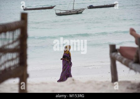 Donna musulmana passeggiate sulla spiaggia di Nungwi, Zanzibar, coperte dalla testa ai piedi e indossa una sciarpa di testa in una giornata di sole in spiaggia, dhows dietro di lei Foto Stock