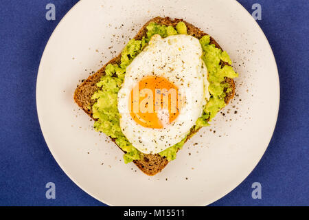 Uovo fritto Sunny Side fino a avocado frantumato e pane di segale aperto di fronte a Sandwich contro uno sfondo blu Foto Stock