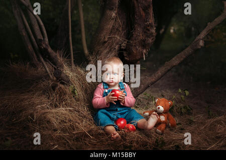 Bambina con un giocattolo orsacchiotto seduto sotto un albero. Lei mangia un Apple Foto Stock