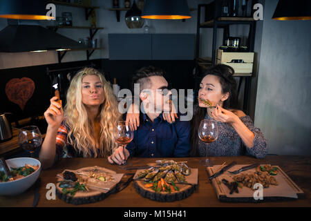 Due giovani donne con un uomo ha la cena insieme. Gruppo di persone interessanti si siede e si mangia il cibo e le bevande vino e godersi il tempo Foto Stock