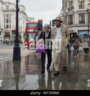 Regent Street, Londra. Una coppia di anziani camminare a braccetto verso il basso di un umido e Rainy Street vicino a Oxford Circus con Bus londinese e dello shopping in background Foto Stock