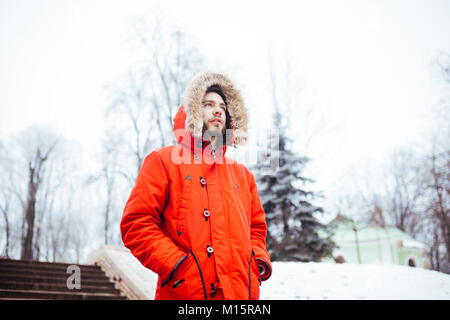 Ritratto di un giovane uomo elegante con la barba vestito di rosso giacca invernale con cappuccio e pelliccia sulla sua testa si erge contro il fondale di una coperta di neve ci Foto Stock