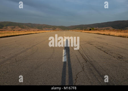 Raggiungere l'orizzonte ampio dritta strada asfaltata, ex striscia aereo al tramonto la lunga ombra del fotografo visto sul vecchio asfalto Foto Stock