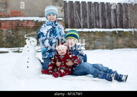 Poco carino sorridente baby boy e i suoi due fratelli più anziani, seduti all'aperto nella neve pupazzo di neve accanto a loro Foto Stock