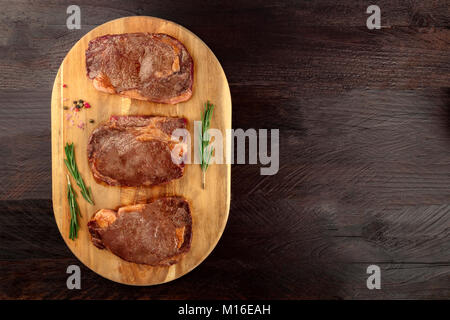 Una foto aerea di tre fette di carne cotta, filetti di manzo, ripresa dall'alto sul buio rustica con rametti di rosmarino, sale, pepe e un p Foto Stock