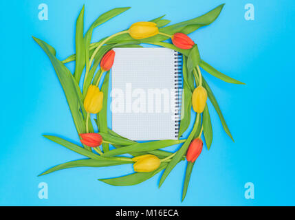 Foglio bianco di notebook circondato da rosso e tulipani gialli cerchio blu su sfondo color pastello. Foto Stock