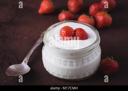 Yogurt bianco in recipiente di vetro con cucchiaio e starwberries su sfondo rustico. Foto Stock