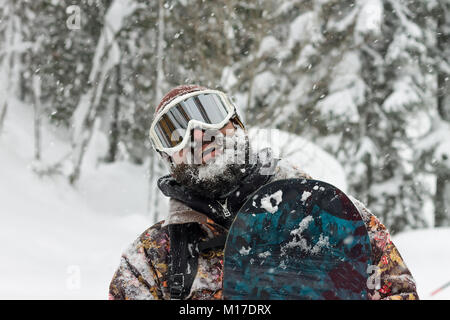 Ritratto di uomo barbuto occhiali da sci snowboard di contenimento in montagna. Foto Stock