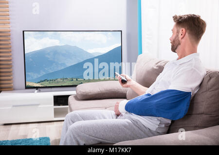 Giovane uomo con mano fratturata seduti sul divano a guardare la televisione a casa Foto Stock