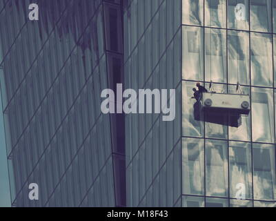 Addetto alla pulizia appeso alla funivia fuori dalla facciata grattacielo con una parete a tenda di vetro che riflette i cieli Foto Stock