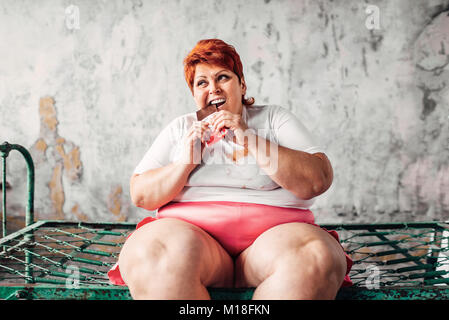 Il sovrappeso donna si siede sul letto e si mangia il cioccolato, la pigrizia e l'obesità, bulimic. Cibo malsano mangiare, femmina grassi Foto Stock