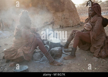 Due hang-ragazze la cottura nella parte anteriore di una capanna di fango dal fuoco,Kaokoveld,Kunene,Namibia Foto Stock