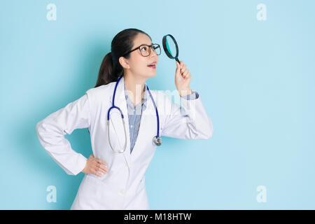 Bellezza sorridente ragazza medico che trasportano uno stetoscopio in piedi di sfondo blu e utilizzando lo strumento Magnifier guardando sopra la ricerca di informazioni mediche. Foto Stock
