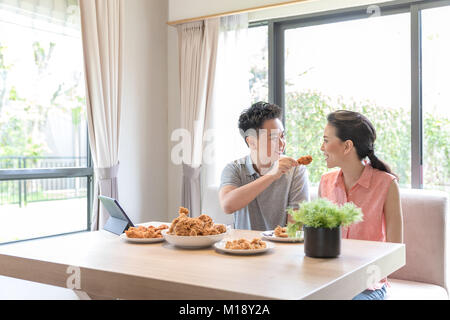 Giovani coppie asiatiche mangiare pollo fritto insieme nel salotto di casa contemporanea per lo stile di vita moderno concetto Foto Stock