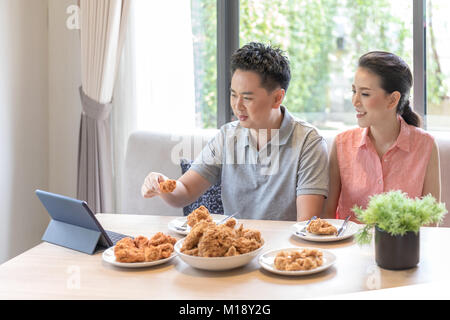 Giovani coppie asiatiche mangiare pollo fritto insieme nel salotto di casa contemporanea per lo stile di vita moderno concetto Foto Stock