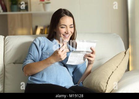 Felice donna incinta la lettura di un foglio illustrativo prima di prendere una pillola seduta su un divano nel salotto di casa Foto Stock