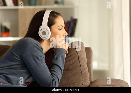 Vista laterale verticale di un triste pensieroso donna ascoltando la musica seduto su un divano nel salotto di casa Foto Stock