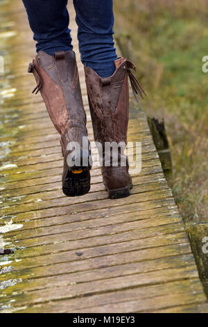 Una donna passeggiate in campagna con terreni fangosi stivali in pelle su una vecchia passerella in legno che indossa jeans e stivali da cavallo in campagna. Nella contea di moda Foto Stock