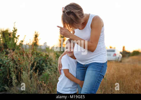 I valori della famiglia. Gravidanza La madre e il figlio sono a piedi nel campo al tramonto. Ella mostra il ragazzo da qualche parte e il Figlio abbraccia la sua gamba. Foto Stock