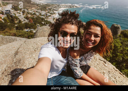 Giovane uomo e donna seduta insieme sulla cima della scogliera prendendo self portrait. Coppia felice seduto sulla cima della montagna e prendendo selfie. Foto Stock