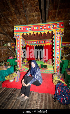 Piuttosto giovane donna malese che pongono di fronte a un raffinato letto in un tradizionale longhouse, Mari Mari Villaggio Culturale, Kota Kinabalu, Sabah Borneo, Ma Foto Stock