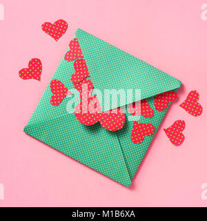 Elevato angolo colpo di alcuni cuori rossi modellato con puntini verdi uscendo da una busta verde modellato con punti gialli, su uno sfondo rosa Foto Stock