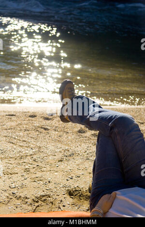 Vista ritagliata della donna gli escursionisti di gambe incrociate di indossare scarpe da trekking come ella si stende su una spiaggia da un fiume Foto Stock