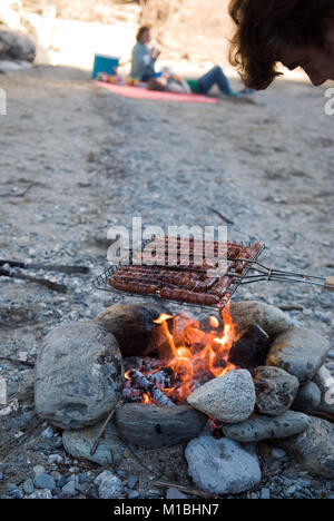 In primo piano un uomo cuochi salsicce su un barbecue in casa fatta con pietre su una spiaggia mentre la gente aspetta un picnic rug in background Foto Stock