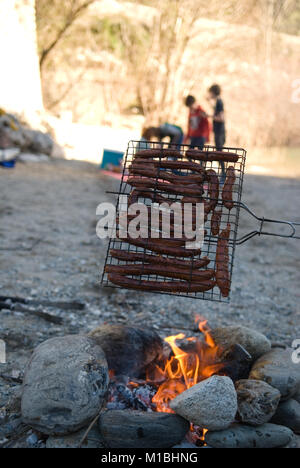 In primo piano qualcuno tiene la cottura di salsicce su un barbecue in casa fatta con pietre su una spiaggia mentre la gente aspetta un picnic rug Foto Stock