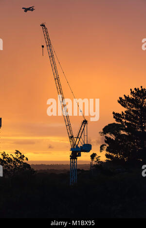 Una costruzione gru su un sito di costruzione con un piano passante per stagliano contro un arancione tramonto di Sydney, Australia Foto Stock