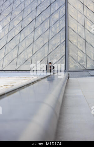 Un uomo si siede sul fondo della famosa piramide di vetro ingresso, guardando il modello geometrico della struttura, il museo del Louvre a Parigi, Francia Foto Stock