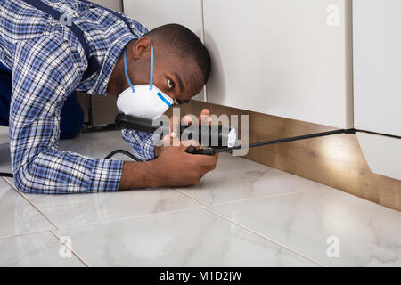 Lavoratore di sesso maschile con maschera inginocchiati sul pavimento e la spruzzatura di pesticidi su cabinet in legno utilizzando la torcia elettrica Foto Stock