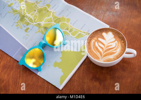 White tazza di caffè con latte art con mappa dei viaggi e gli occhiali da sole sul legno marrone tabella,le attività per il tempo libero. Foto Stock