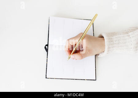 Close-up di mani femminili in maglione azienda golden balpoint penna e scrittura su notebook aperto sul tavolo bianco sullo sfondo. L'istruzione e il concetto di pianificazione. Stile immagine di stock. Foto Stock