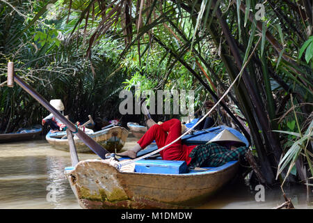 Rematore appoggiata in barca nel delta del Mekong, Vietnam. Rosso brillante pantaloni forniscono grande contrasto nel selvaggio della giungla verde Foto Stock