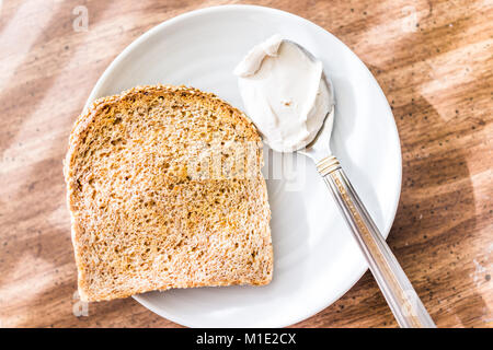 Primo piano della fetta pezzo di tutto il grano germogliato tostati Pane di grano sulla piastra, cucchiaio con il bianco crema di formaggio sulla tavola piana, vista dall'alto in basso macro Foto Stock