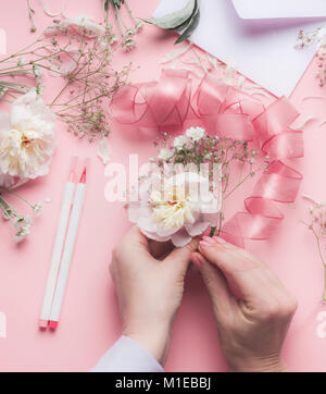 Mani femminili rendendo composizioni floreali con fiori di colore bianco e il nastro di inchiostro a colore rosa pastello sfondo, vista dall'alto Foto Stock