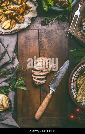 Fette di maiale fritto carne e coltello da cucina sul tagliere con patate al forno sul tavolo rustico, vista dall'alto Foto Stock