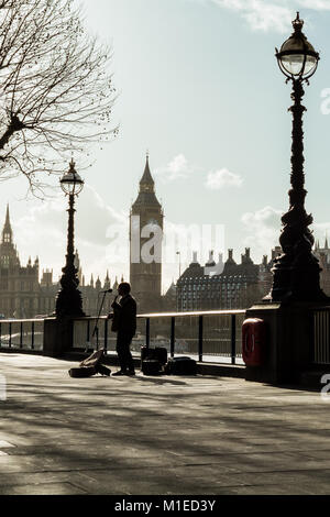 Retro illuminato e stagliano dal tardo pomeriggio di sole invernale, busker gioca sul London South Bank, con Big Ben in background Foto Stock